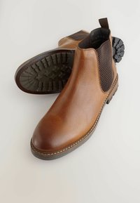 Barna bőr Chelsea csizmák, textúrált elasztikus oldalsó panelekkel és strapabíró gumitalppal, sima világos háttér előtt.