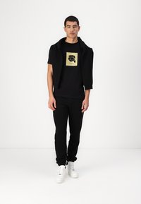 Schwarzes T-Shirt mit kurzen Ärmeln und goldener Grafikdesign, kombiniert mit schlanken schwarzen Jogginghosen und weißen Turnschuhen; lässige schwarze Jacke über den Schultern getragen.