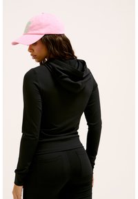 Haut noir à manches longues avec capuche, design ajusté, associé à une casquette rose portant des lettrages verts. Tissu lisse et détails de couture soignés.