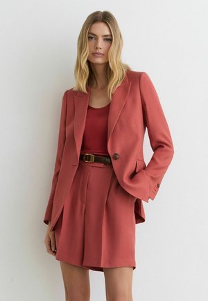 Vrouw met blond haar draagt een roestkleurige blazer en bijpassende geplooide shorts over een rode top, staand tegen een witte muur.