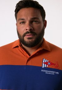 Polohemd in Orange und Blau, mit einem gestickten, meeresbezogenen Logo, das ein Segelboot und Flaggen zeigt. Heller Stoff mit Kragen und Knöpfen.