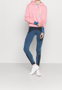 Rosa sweatshirt med huva och färggược logo, i kombination med blå mönstrade leggings med svarta detaljer och vita sportskor.