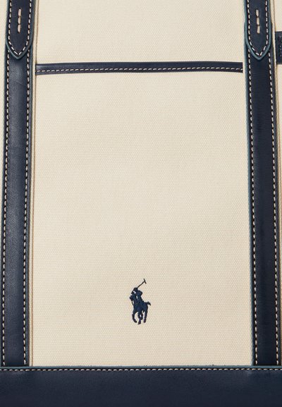 Beige lærredstaske med marineblåt læderkant og syninger, med en lille marineblå polospiller-logo i midten.
