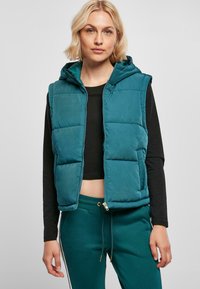 Gilet rembourré teal avec capuche, fermeture éclair et motif matelassé. Porté par-dessus un haut noir, associé à un pantalon de jogging vert. Texture douce.