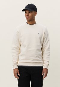 Sweat-shirt beige avec poignets côtelés et col rond, arborant un petit logo bleu foncé. Assorti à une casquette noire et un jean foncé.