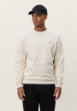 Μπεζ φούτερ με ριμπ μανσέτες και στρογγυλή λαιμόκοψη, διακοσμημένο με μικρό σκούρο μπλε λογότυπο. Συνδυασμένο με μαύρο καπέλο και σκούρο τζιν.