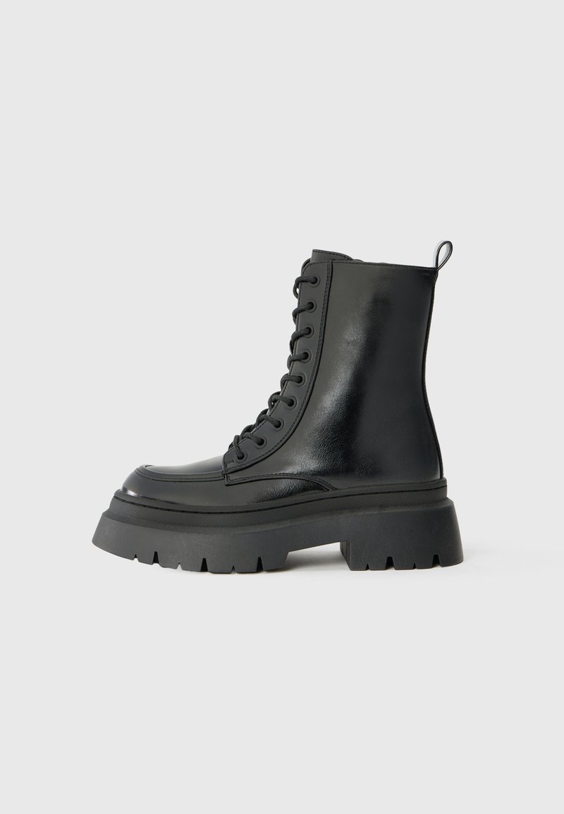 Bottes chelsea noires en cuir à lacets avec une semelle épaisse, présentant une finition brillante, un renfort à l'avant et des crampons prononcés pour une traction optimale.