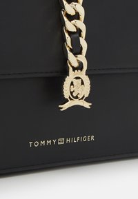 Bolso de cuero negro con una correa de cadena dorada y un emblema con un león. Texto del logo en dorado "TOMMY HILFIGER" grabado en la parte inferior.