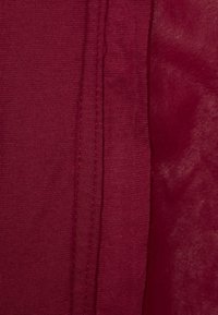 Bordeauxfarbener Stoff mit glatter Textur, der eine Seitennaht-Detail aufweist. Das Material wirkt weich und an einigen Stellen leicht durchsichtig.