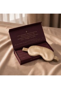 Zijden beige oogmasker met elastisch bandje liggend op een open donkerpaarse doos met gouden tekst op een beige stoffen oppervlak.