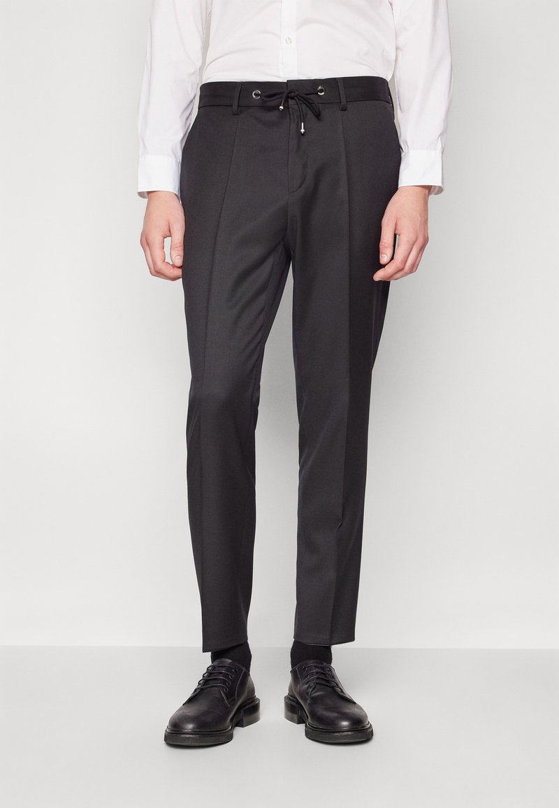 Pantaloni negri, slim-fit, cu talie ajustabilă, pliuri frontale și textură netedă, asortați cu pantofi negri și o cămașă albă.