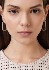 Boucles d'oreilles en or en forme rectangulaire, orn�ées de strass transparents. Haut tissé blanc texturé. Peau de ton lisse.