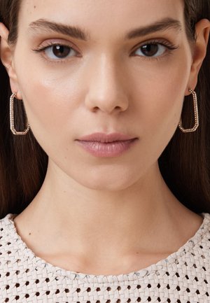 Boucles d'oreilles en or en forme rectangulaire, ornées de strass transparents. Haut tissé blanc texturé. Peau de ton lisse.