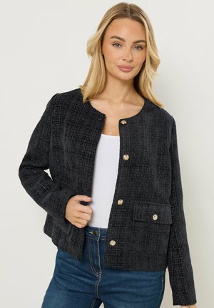 Blondynka w czarnej, teksturowanej kurtce na guziki, na białym topie i niebieskich dżinsach, stojąca na tle jasnego, jednolitego tła.