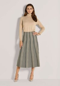 Frau in beigem Rollkragenpullover, schwarz-weißer vertikal gemusterter Midirock und beigen High Heels, steht vor einer hellen Wand.