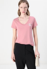Roze V-hals t-shirt van zacht materiaal, met een klein wit logo op de linkerkant van de borst. Gecombineerd met zwarte broeken en een lichtkleurige clutch.