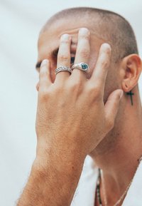 Silverringar på en hand, en med en turkos sten och en annan med ett mönstrat design. Hudens textur och en korsörhänge synliga.