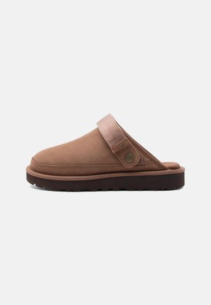 Zoccolo marrone senza lacci con cinturino in tessuto regolabile e bottone in metallo, caratterizzato da una suola nera spessa e robusta adatta all'uso all'aperto.