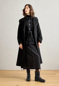 Palton lung din lână neagră peste un ansamblu stratificat din denim negru, având o talie evidențiată prin curea, și ghete din piele neagră cu talpa texturată.