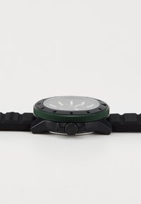 Montre noire avec un cadran vert, bracelet en caoutchouc et face transparente. Présente des marqueurs d'heure blancs et une couronne sur le côté pour les réglages.