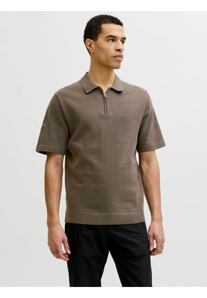 Homme debout, portant un polo marron à manches courtes avec un col zippé et un pantalon noir, regardant légèrement vers la droite.