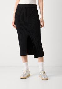 Jupe midi noire en maille tricotée avec une texture côtelée et un motif de tricot en câble, dotée d'une fente avant. Associée à des baskets en argent et des chaussettes blanches.