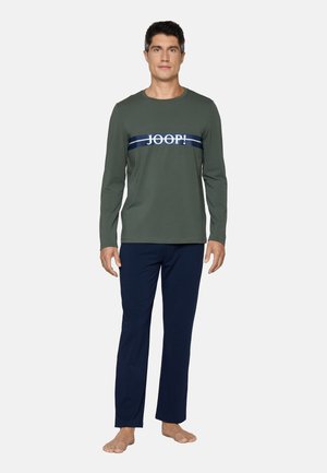 Man staat blootsvoets, draagt een olijfgroen shirt met lange mouwen met tekst "JOOP!" en donkerblauwe broek op een effen achtergrond.