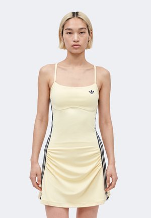 Joven con pelo rubio corto que lleva un vestido Adidas amarillo claro con tirantes finos y rayas laterales negras, de pie mirando hacia adelante.