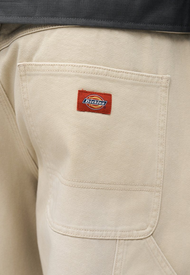 Pantalon beige avec une poche arrière carrée comportant une petite étiquette de marque Dickies rouge et bleue cousue près du centre supérieur.