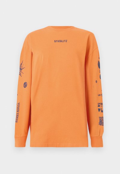 Narancssárga, hosszú ujjú pamut póló. Az elején a "STIEGLITZ" felirat található, míg az ujjak különböző kék grafikai mintákkal díszítettek.
