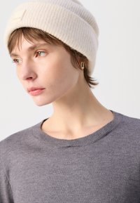 Cremefarbene Rippmütze, grauer Strickpullover mit rundem Ausschnitt. Goldene geometrische Ohrringe. Weiche Texturen, neutrale Farben und modernes Design.