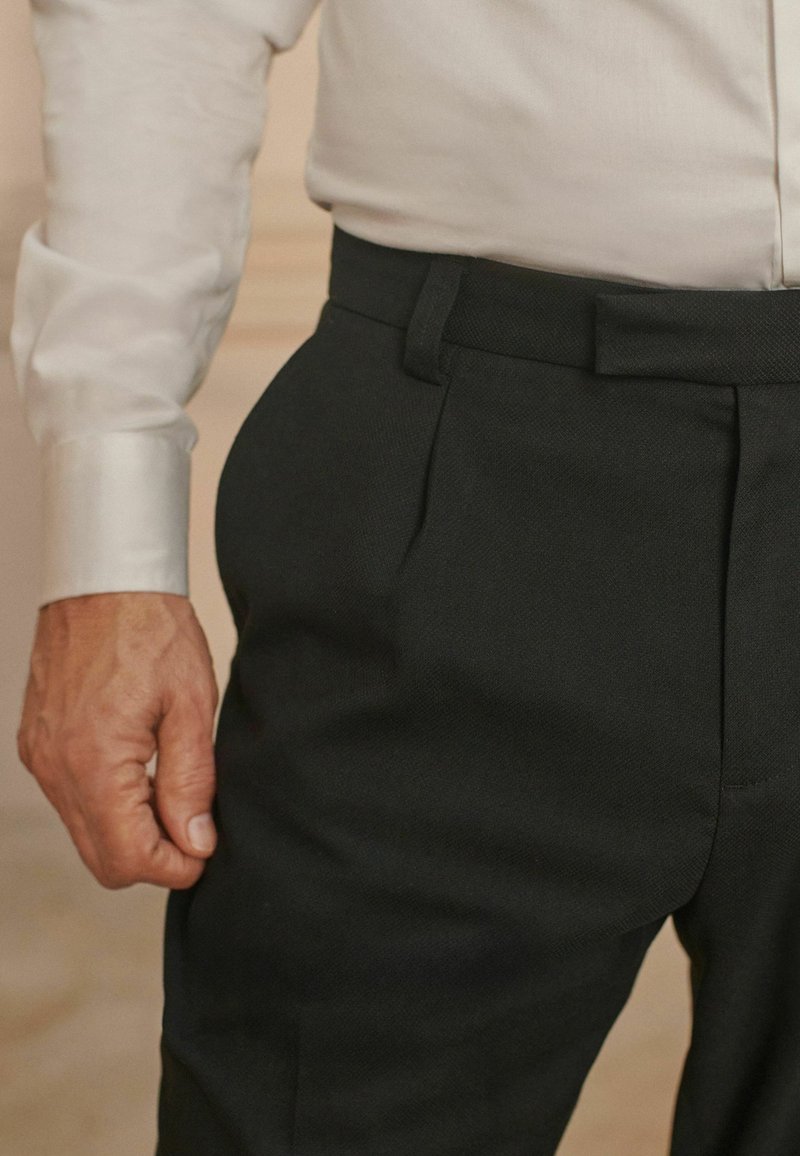 Schwarze, strukturierte Hose mit Seitentaschen, flachem Bündchen und Gürtelschlaufen. Dazu passt ein weißes Hemd mit umgeschlagenen Ärmel.