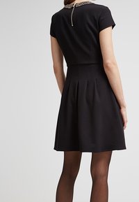 Schwarzes Kleid mit kurzen Ärmeln, tailliertem Oberteil, gefaltetem Rock und dekorativem Kragen. Hat einen Reißverschluss am Rücken. Glatte Stofftextur.