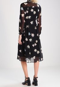 Svart blommig klänning med långa genomskinliga ärmar. Har en figursydd överdel och en flödande kjol, prydd med pastellfärgade blommönster. Svarta klackskor.