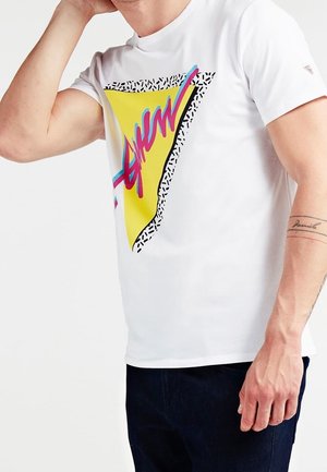 T-shirt blanc à manches courtes avec un grand motif en forme de triangle jaune, rehaussé d'accents roses et bleus, bordé d'un motif à pois noir et blanc.
