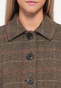Veste en pied-de-poule brun avec un col classique, deux grands boutons noirs et des accents à carreaux rouges et bleus sur un tissu texturé.