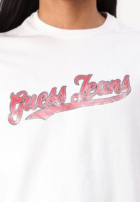 Hvit bomulls t-skjorte med en stor, glitrende rød og svart "Guess Jeans"-logo i kursiv stil over forsiden.