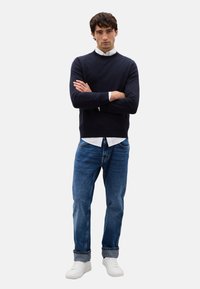 Maglione blu navy, camicia bianca con colletto sotto, jeans in denim blu e sneakers bianche. Il modello sta in piedi con le braccia incrociate, su uno sfondo neutro.