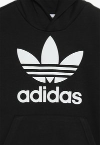 Mustvalge kapuutsiga dressipluus, mis on valmistatud puuvilla segu materjalist, esiküljel on suur valge Adidas trefoil logo ja tekst, lisaks on ees kenguru tasku.