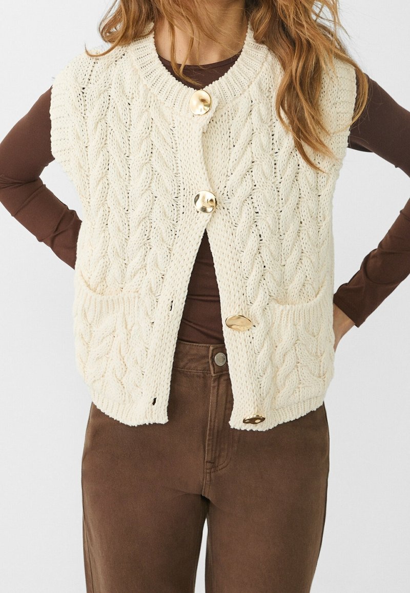 Crème kleurige kabelgebreide mouwloze vest met grote gouden knopen, gedragen over een bruine lange mouwen top en bruine broek.