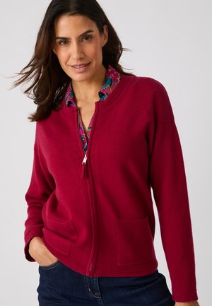 Mujer que lleva un suéter rojo con cremallera sobre una camisa floral, con su mano izquierda en el bolsillo, sonriendo suavemente.