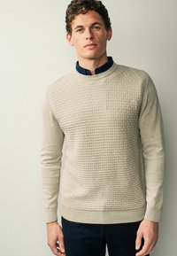 Pull en beige texturé avec un motif tissé, col rond et poignets côtelés. Superposé sur une chemise à col bleu marine.