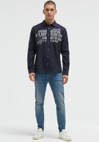 Dunkelblaues Langarmhemd mit weißen, von der Luftfahrt inspirierten Grafiken, Knopfleistenkragen und strukturiertem Stoff, kombiniert mit hellblauen Jeans und weißen Sneakers.