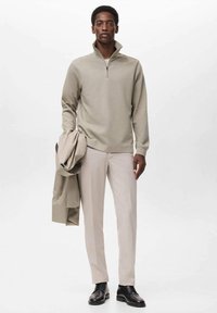 Pulóver de cuello con cremallera en gris claro, hecho de un tejido suave, combinado con pantalones beige y zapatos negros. El modelo sostiene una chaqueta beige bajo un brazo.