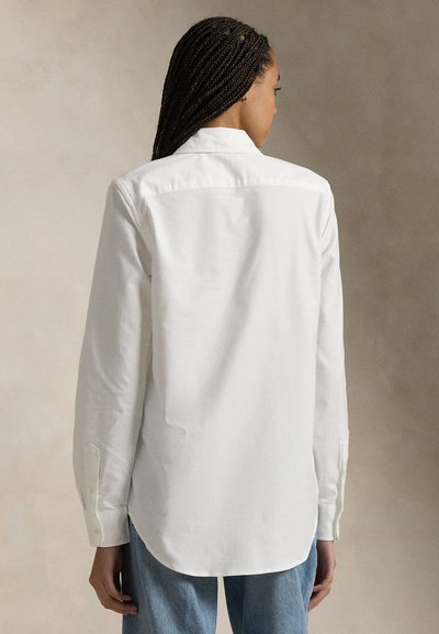 Kvinne med fletter som har på seg en hvit langermet skjorte og blå jeans, vist bakfra mot en nøytral bakgrunn.