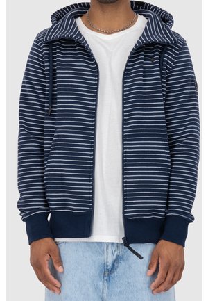 Mand iført en navyblå og hvidstribet lynlåshættetrøje over en hvid t-shirt, kombineret med lyseblå jeans og en sølvfarvet kædehalskæde.