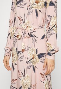 Abito floreale con maniche lunghe, tessuto rosa chiaro, vita con cintura, bottoni lungo la parte anteriore, con grandi fiori color crema e blu e vegetazione.