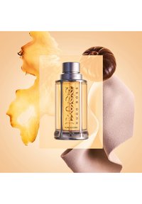 Hugo Bossin tuoksupullo, jossa on kirkas lasikapseli, hopeinen kansi ja kultainen etiketti; ympäröity inkiväärillä, kastanjalla ja lämpimissä sävyissä olevalla teksturoitulla kankaalla.