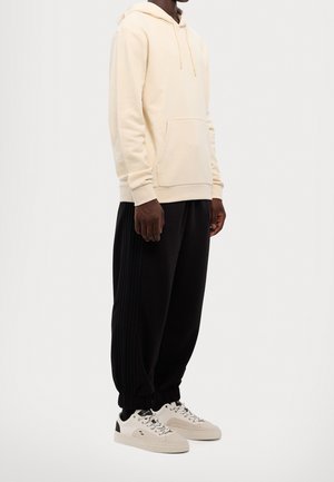 Person trägt cremefarbenen Hoodie, schwarze Jogginghose mit gerippten Seitenteilen und beigefarbene Sneakers mit weißen Schnürsenkeln und steht vor weißem Hintergrund.