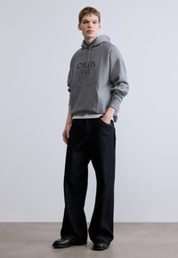 Szürke pamut kapucnis pulóver első zsebbel és logónyomattal, sötét farmer bő szárú nadrággal és fekete lábbelivel párosítva. Egyszerű, hétköznapi design.